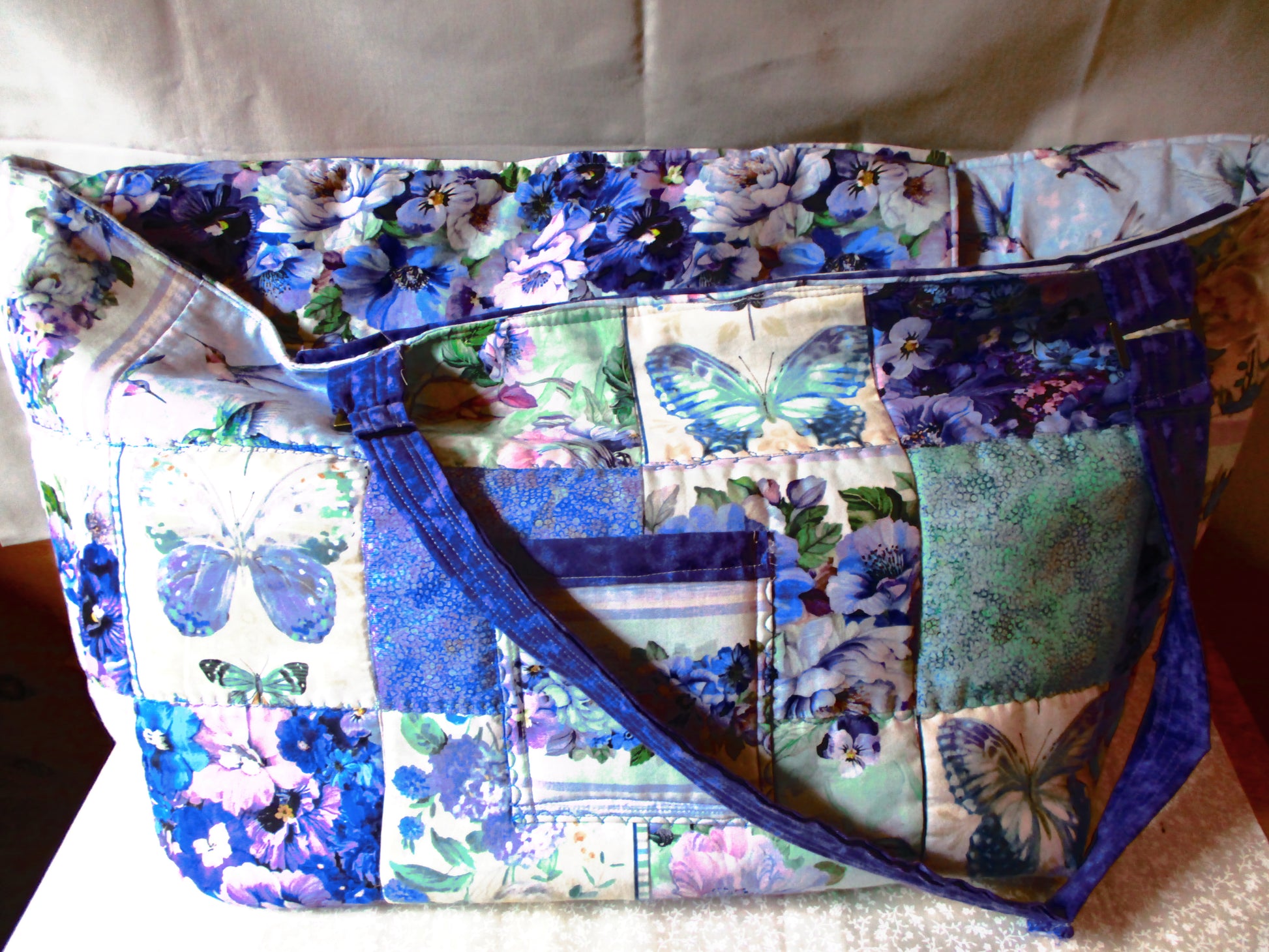 A patchwork quilted tote bag featuring blue and purple floral prints, butterflies, and solid blue fabric, with a matching blue shoulder strap, set against a neutral fabric backdrop.
