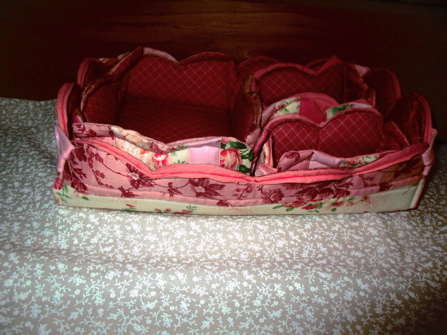 A pink floral fabric organizer tray with multiple compartments, photographed on a patterned surface against a dark wooden background.